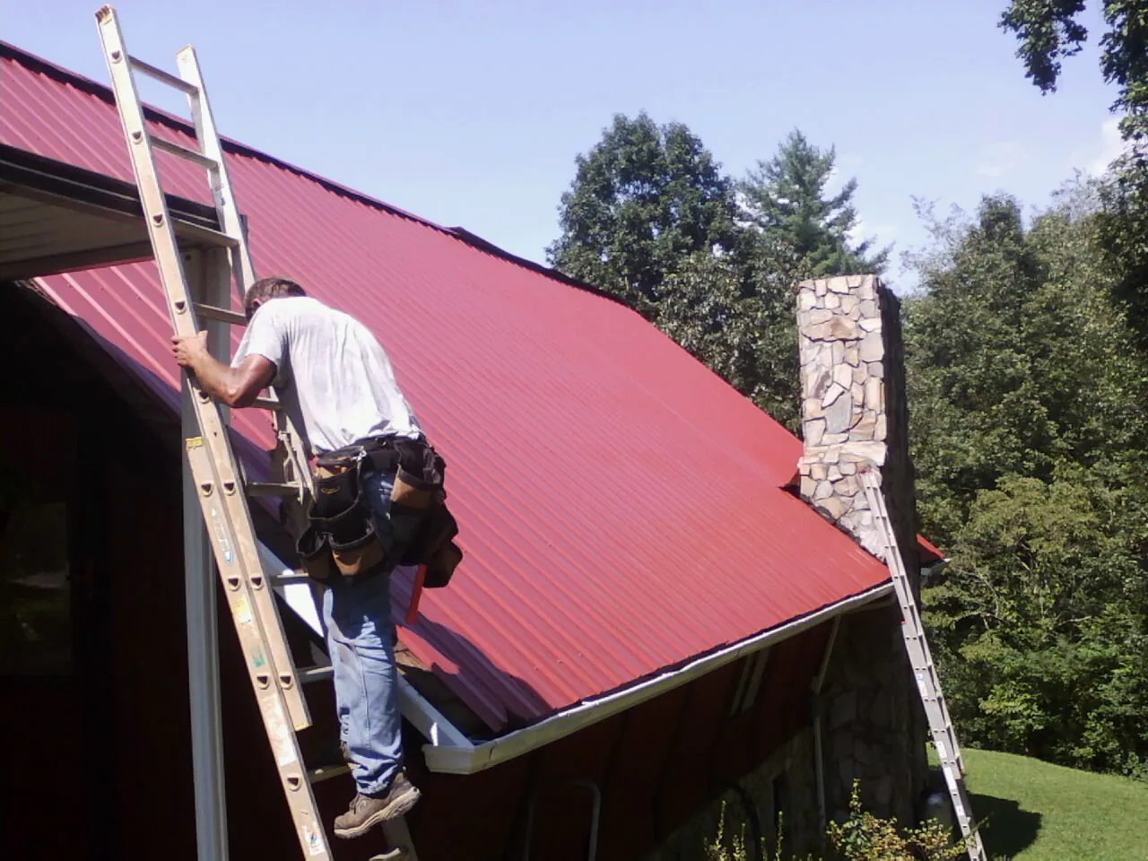 Roofer on a ladder beside a red metal roof and stone chimney — Soffit & Fascia Repair by Roofing Brothers in Saltville